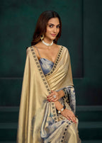 A woman wearing a draped beige and light blue saree with black patterned borders, styled with a matching blue blouse, pearl necklace, and gold jewelry, posed against a dark green background.