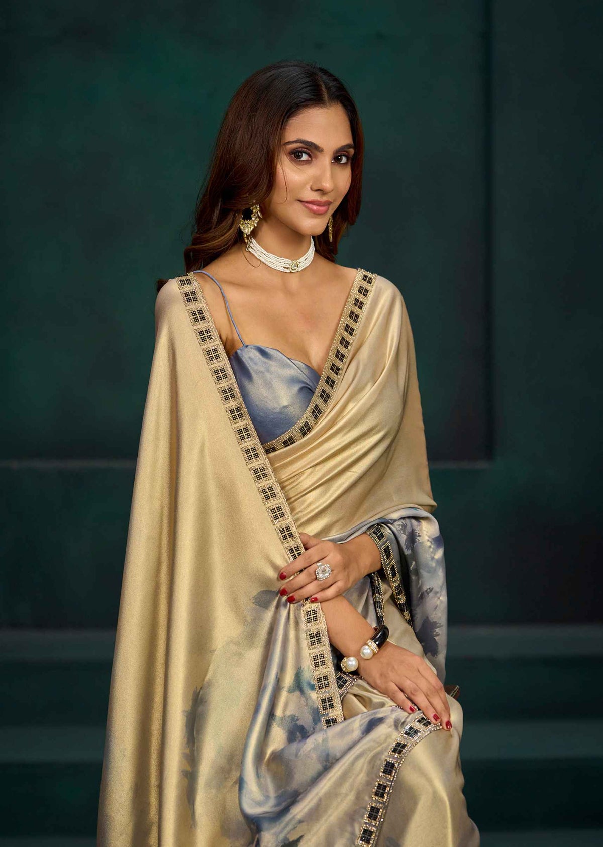 A woman wearing a draped beige and light blue saree with black patterned borders, styled with a matching blue blouse, pearl necklace, and gold jewelry, posed against a dark green background.
