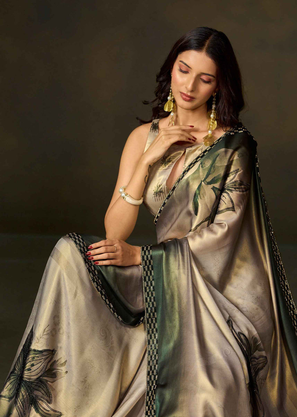 A woman wearing a metallic champagne-gold saree with green and black floral motifs and a patterned border, posed against a dark brown studio background.
