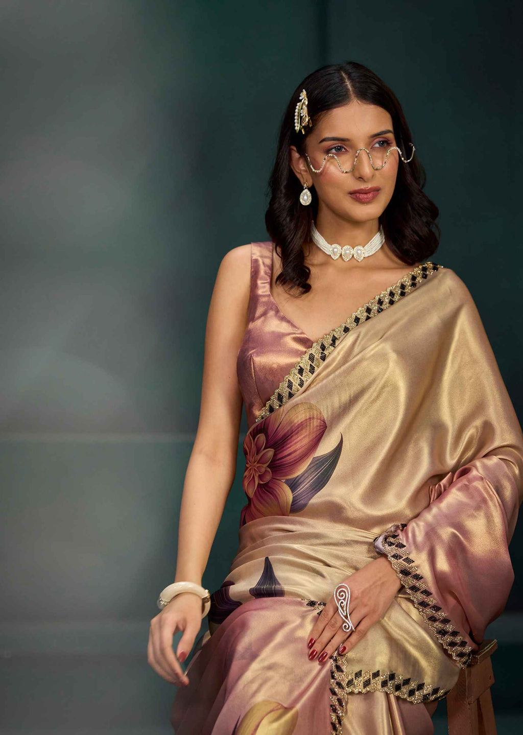A woman models a metallic champagne-gold satin saree with a pink floral print and black geometric border trim, styled with a white collar necklace, gold earrings, and rings against a dark green background.