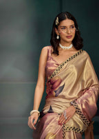 A woman models a metallic champagne-gold satin saree with a pink floral print and black geometric border trim, styled with a white collar necklace, gold earrings, and rings against a dark green background.