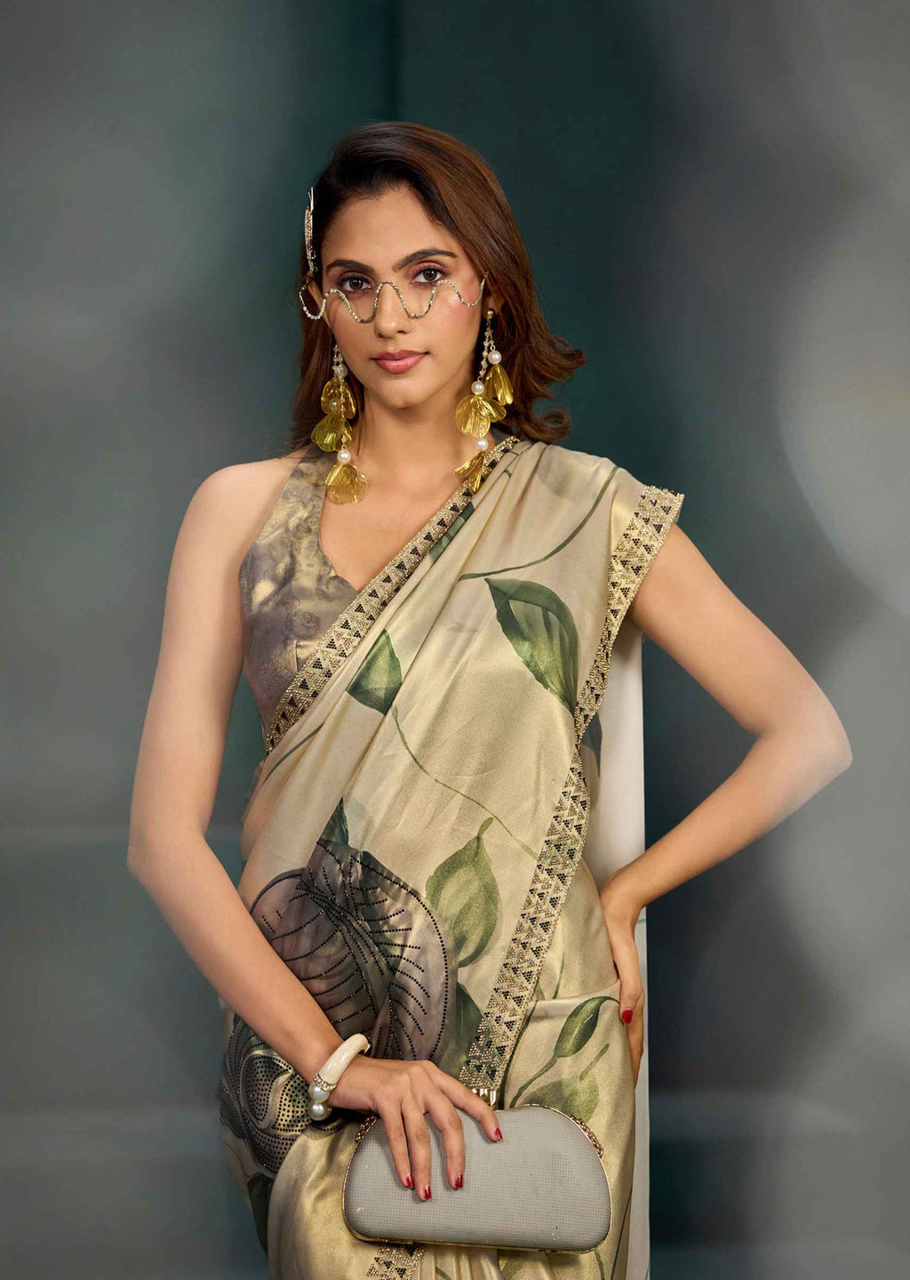 A woman models a beige and green printed saree with a matching sleeveless blouse. She is wearing round wire-frame glasses, gold earrings, and a white bracelet, and holds a gray clutch purse against a dark gray studio background.