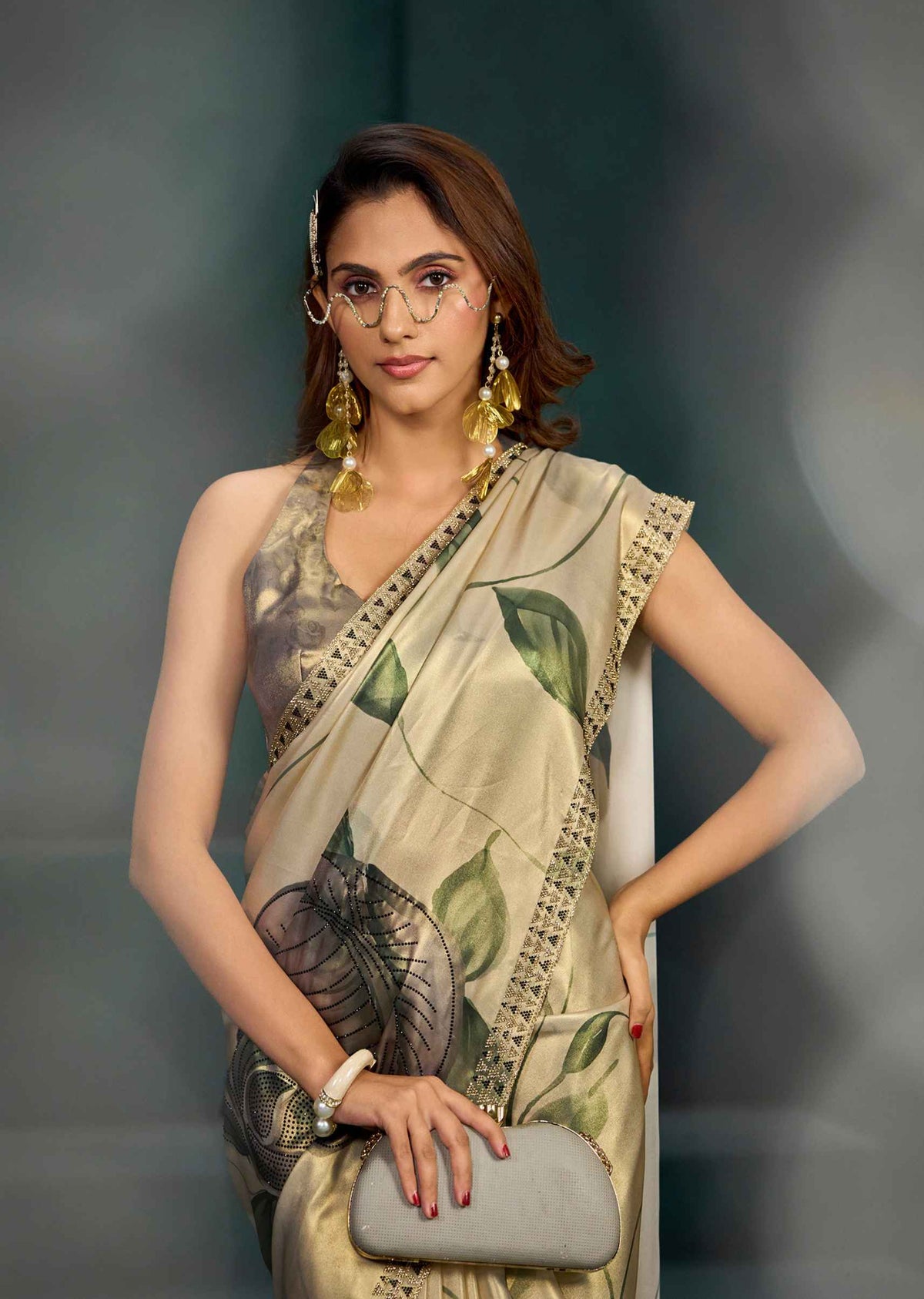 A woman models a beige and green printed saree with a matching sleeveless blouse. She is wearing round wire-frame glasses, gold earrings, and a white bracelet, and holds a gray clutch purse against a dark gray studio background.