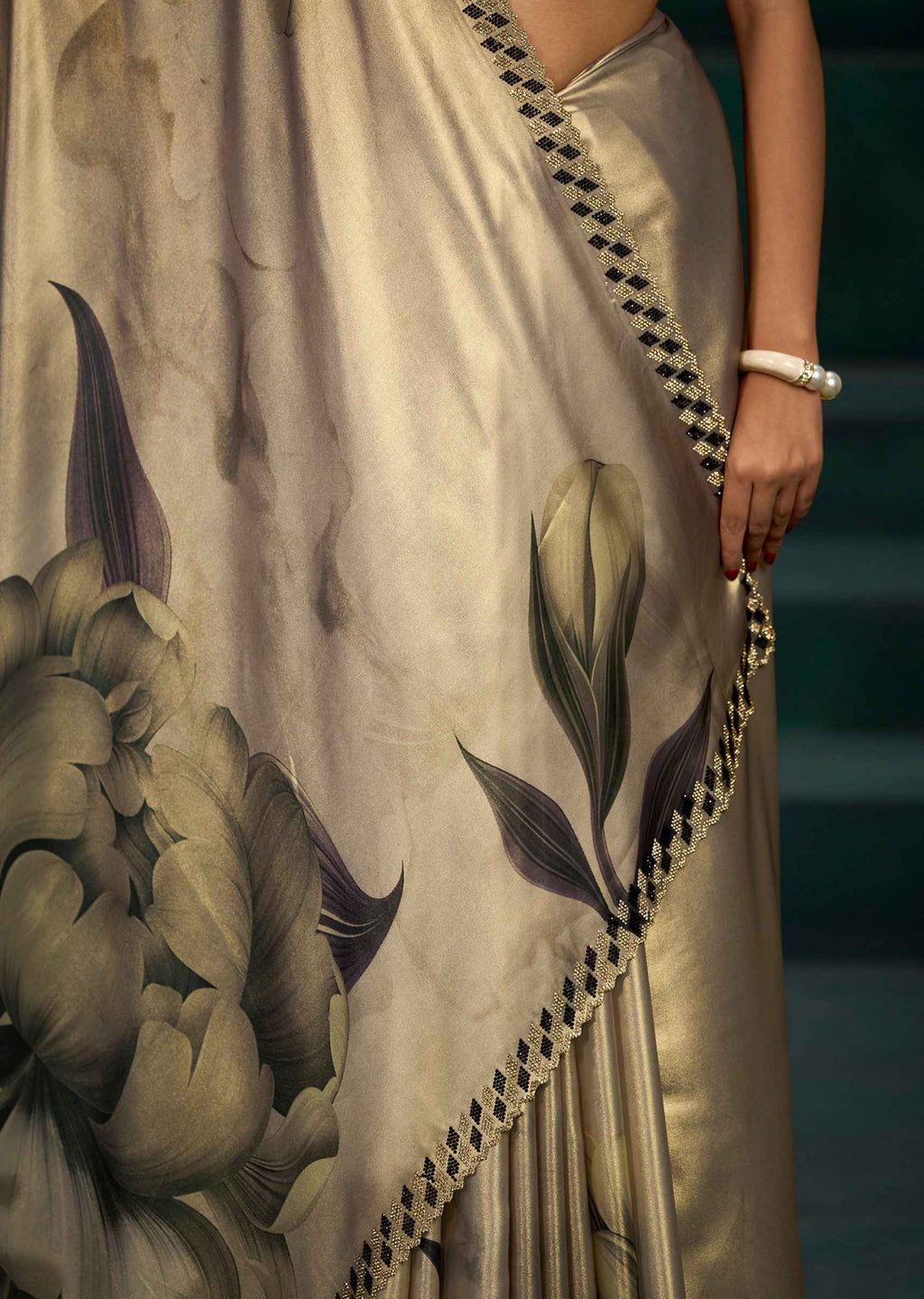 A close-up of a model wearing a beige silk saree with a large floral print in muted green and gray, featuring a black and white checkered border.