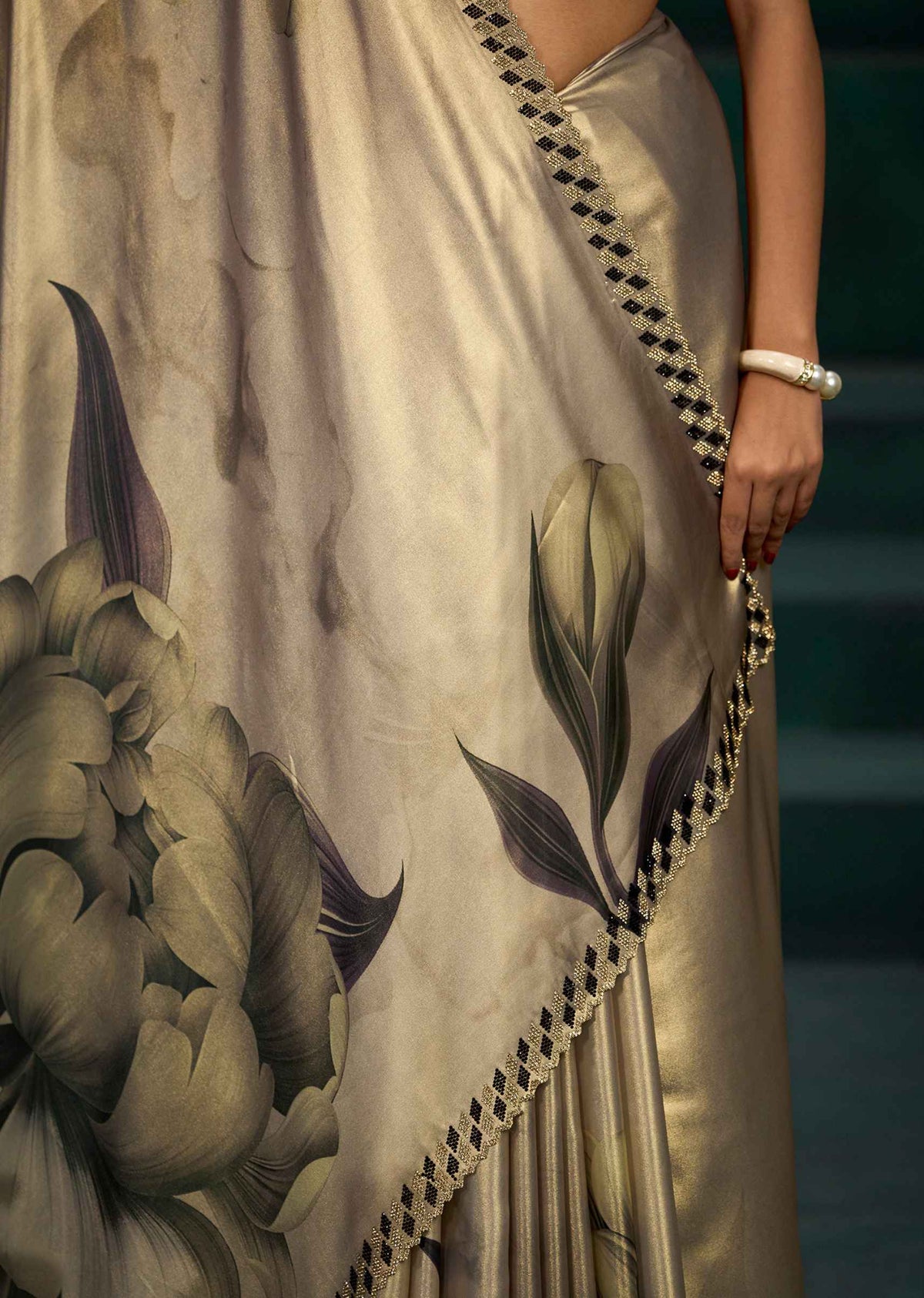 A close-up of a model wearing a beige silk saree with a large floral print in muted green and gray, featuring a black and white checkered border.