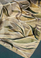 A close-up of a lightweight scarf or fabric with a flowing leaf pattern in olive green, beige, and gold tones, finished with a fringed edge.