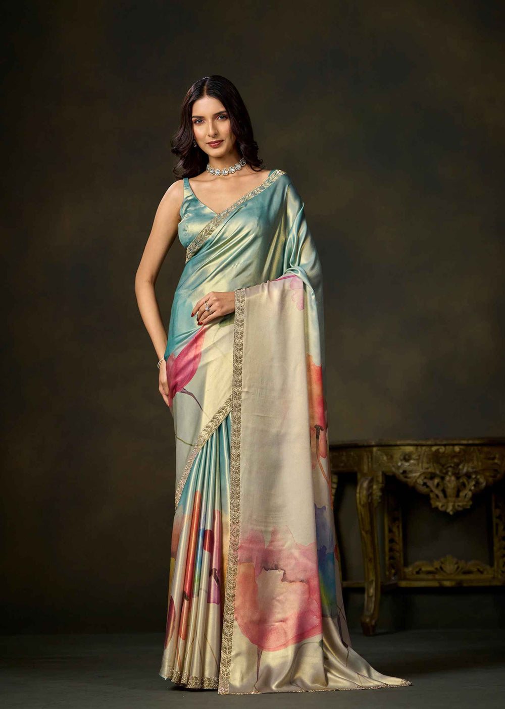 A woman models a draped saree in a studio setting with a dark brown backdrop and an ornate table. The saree has a light blue-green body with a cream border and pink and blue abstract floral patterns.
