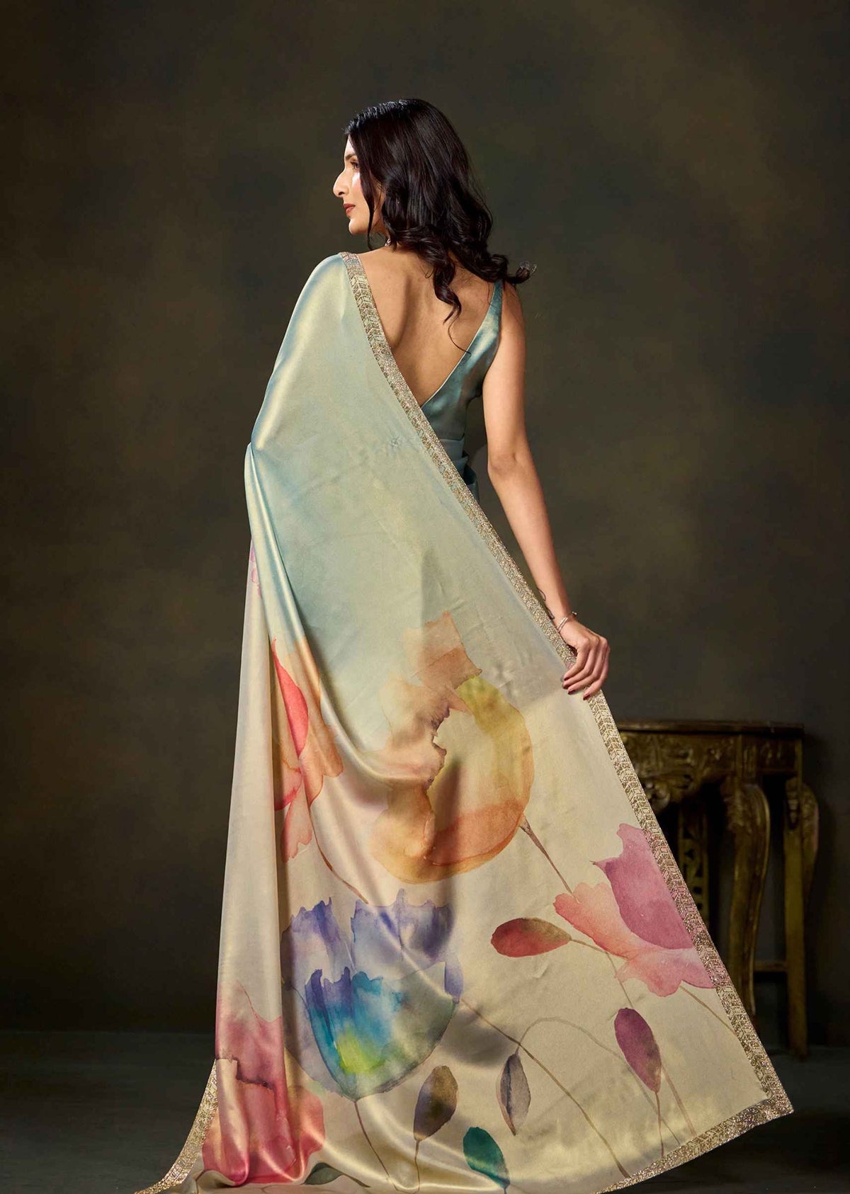 A woman wearing a flowing satin saree with a watercolor floral print in pastel shades of pink, blue, yellow, and green, posed against a dark brown studio background.